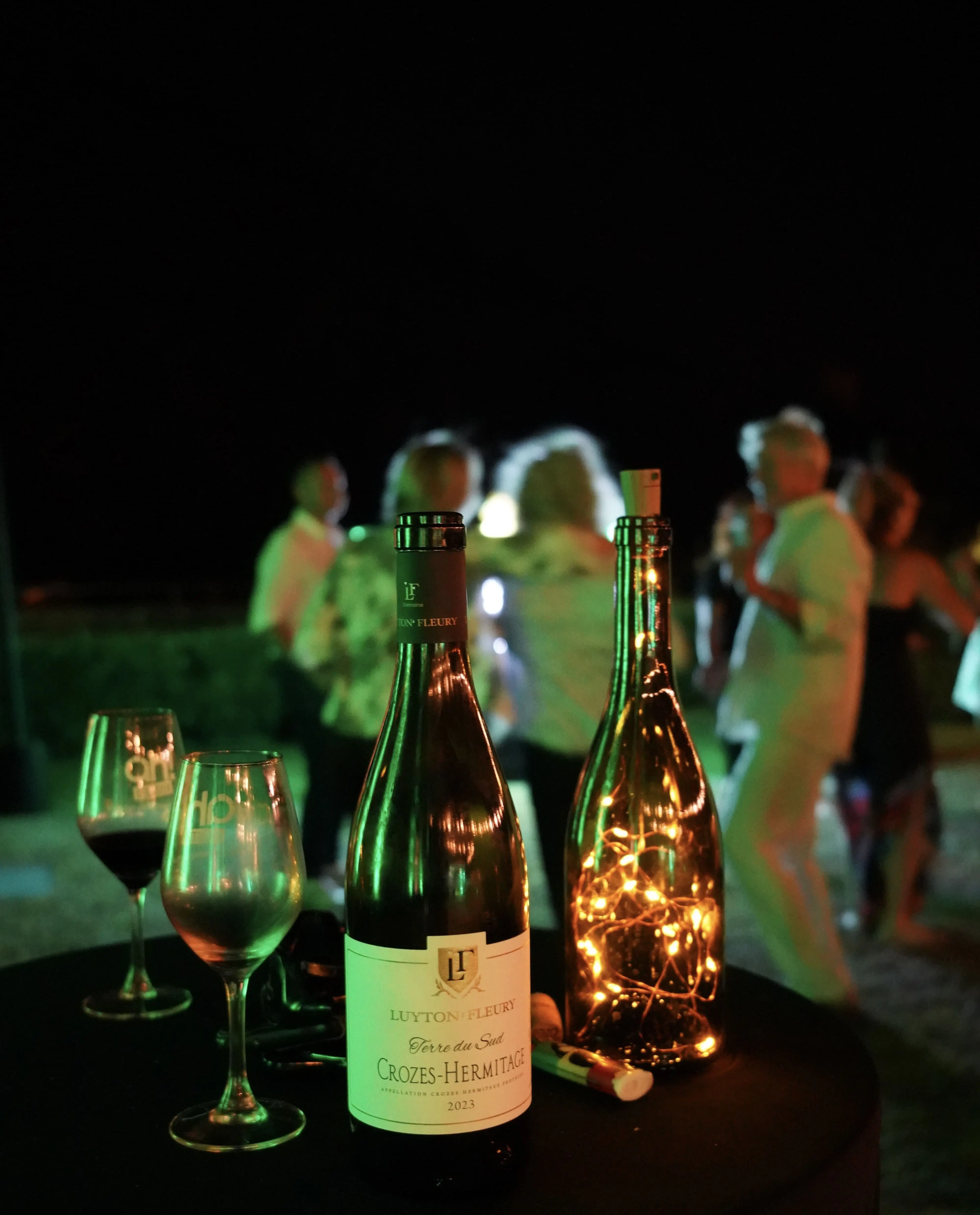 Bouteille de vin Crozes-Hermitage sur table de soirée avec ambiance lumineuse et invités en arrière-plan – photographie événementielle Focus&Toit.